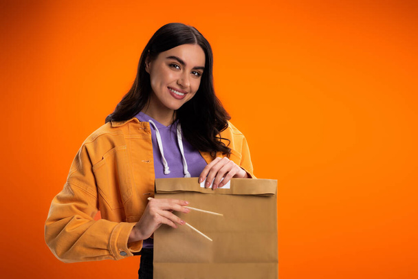 Smiling brunette woman holding bamboo chopsticks and paper bag isolated on orange - Фото, изображение