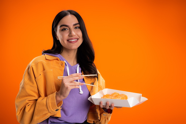 Stylish brunette woman looking at camera while holding chopsticks and sushi isolated on orange - Photo, Image