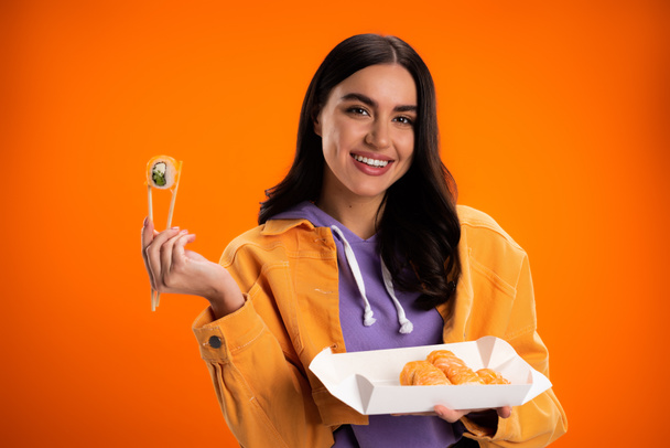 Smiling brunette woman holding sushi and looking at camera isolated on orange - Zdjęcie, obraz