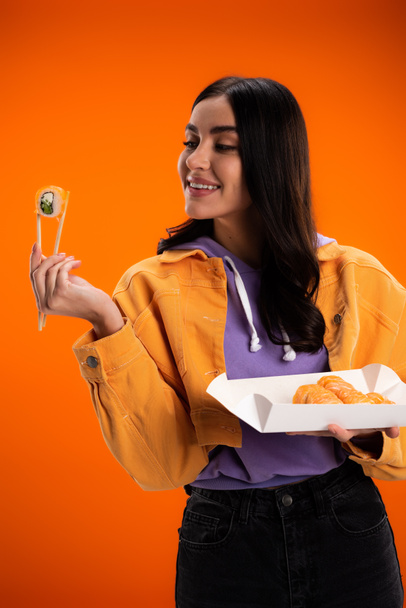 Stylish young woman looking at sushi and chopsticks isolated on orange - Zdjęcie, obraz