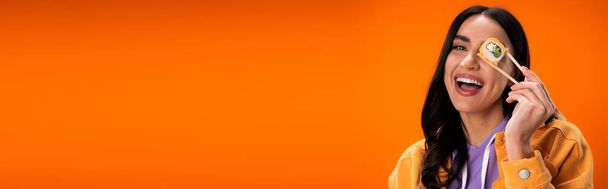 Young woman holding chopsticks and tasty sushi near face isolated on orange, banner  - Photo, Image