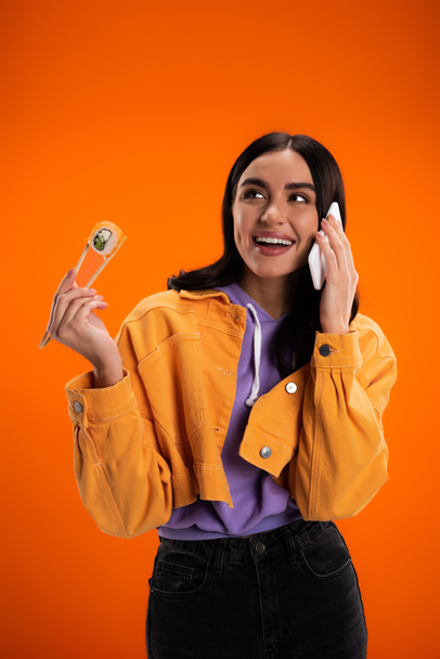 Положительная брюнетка женщина держит суши в палочках и разговаривает на смартфоне изолированы на оранжевый - Фото, изображение