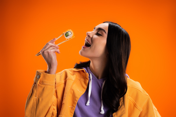 Stylish brunette woman opening mouth and holding sushi isolated on orange - Foto, immagini