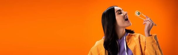 若い女性が口を開けて、オレンジに孤立した箸で寿司を握る、バナー  - 写真・画像