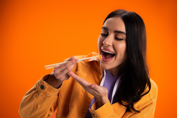 brunette woman in stylish jacket eating delicious sashimi isolated on orange - Foto, Imagen