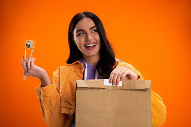 joyful woman holding paper bag and chopsticks with sushi roll while looking away isolated on orange - Foto, Imagem