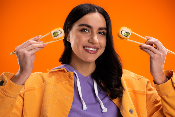 happy brunette woman in jacket and purple hoodie posing with delicious sushi rolls isolated on orange - Foto, immagini