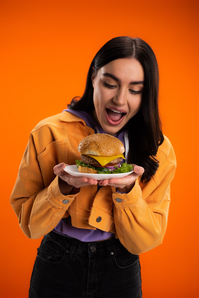 hungry woman opening mouth near plate with delicious burger isolated on orange - Photo, Image