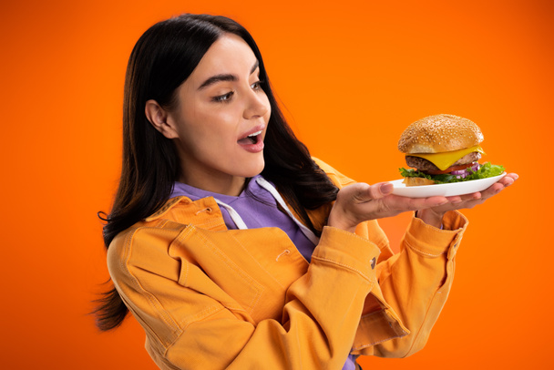  amazed woman in stylish jacket looking at tasty burger with cheese and meat isolated on orange - Photo, Image