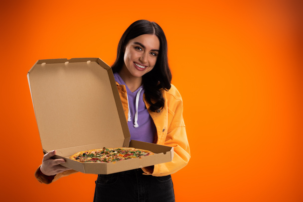 joyful woman looking at camera while holding carton box with tasty pizza isolated on orange - Фото, изображение