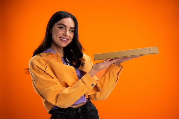 весела жінка в модній куртці тримає коробку для піци і дивиться на камеру ізольовано на помаранчевому
 - Фото, зображення