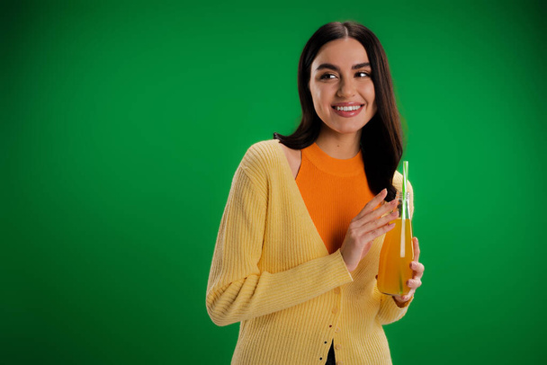 стильная женщина с бутылкой свежего сока улыбаясь и глядя в сторону изолированы на зеленый - Фото, изображение