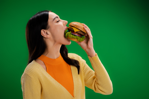 hungry brunette woman eating delicious burger with meat and cheese isolated on green - Foto, Imagem