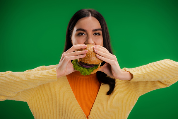 young brunette woman eating delicious burger and looking at camera isolated on green - Photo, Image