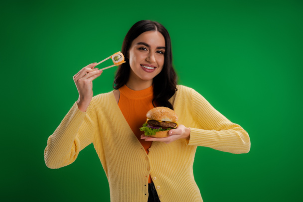 happy young woman holding burger and chopsticks with sushi roll isolated on green - Foto, immagini