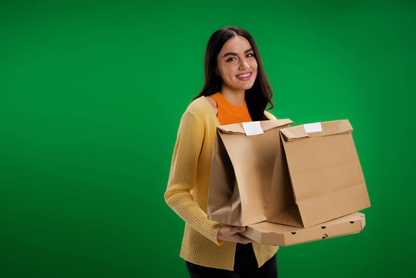 happy brunette woman holding paper bags and pizza box while looking at camera isolated on green - Foto, afbeelding