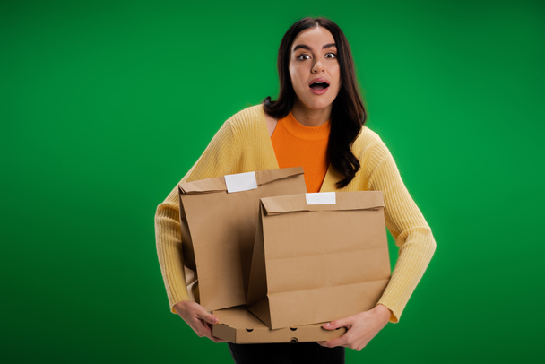 amazed woman with food packages looking at camera isolated on green - Photo, image