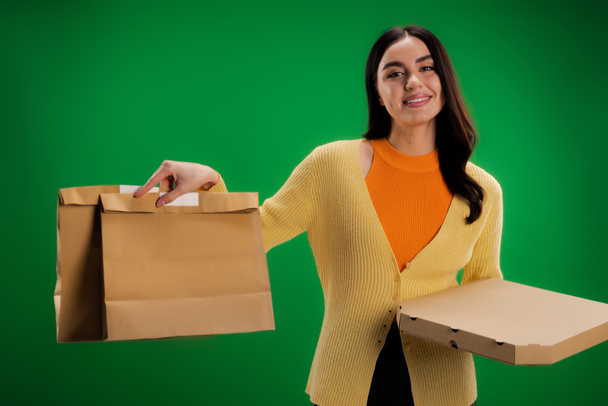 smiling woman holding craft paper bags and pizza box while looking at camera isolated on green - Fotografie, Obrázek