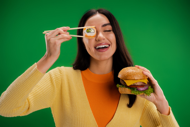 cheerful woman with tasty burger holding sushi roll near eye isolated on green - Photo, Image