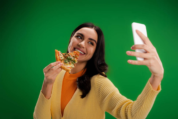 iloinen nainen puree pala pizzaa ja ottaa selfie älypuhelimeen eristetty vihreä - Valokuva, kuva