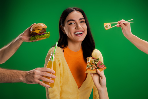 happy young woman looking at camera near people proposing different food isolated on green - Foto, afbeelding