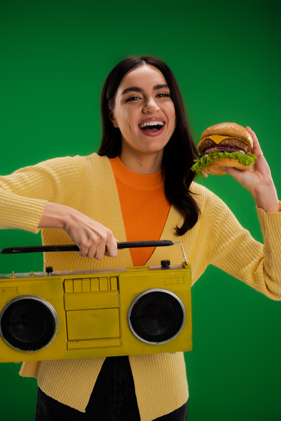 laughing woman holding tasty burger and boombox while looking at camera isolated on green - Photo, Image