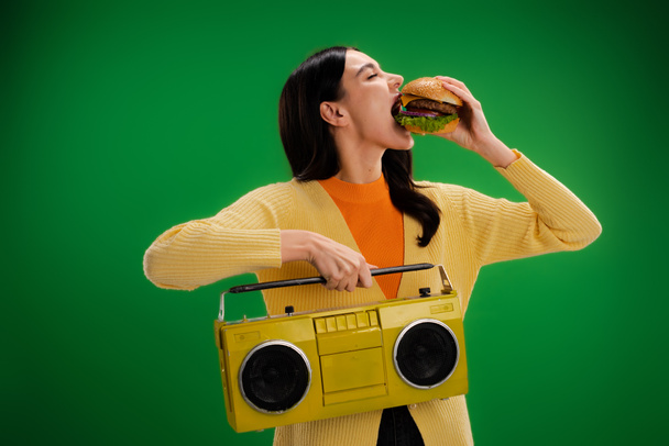 young and hungry woman with boombox eating tasty burger isolated on green - Foto, imagen