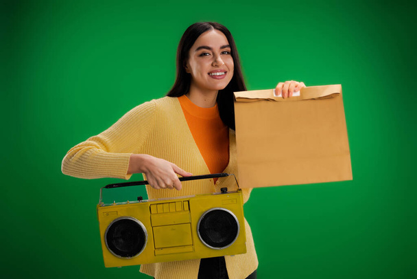 счастливая женщина держит бумбокс и продовольственный пакет изолированы на зеленый - Фото, изображение
