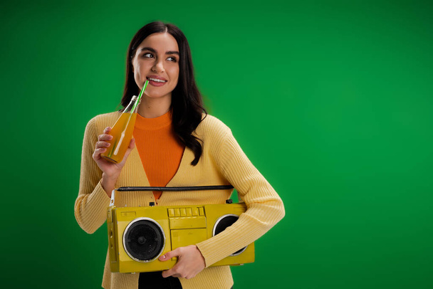 улыбающаяся женщина с бумбоксом пьет газировку и смотрит в сторону изолированы на зеленый - Фото, изображение