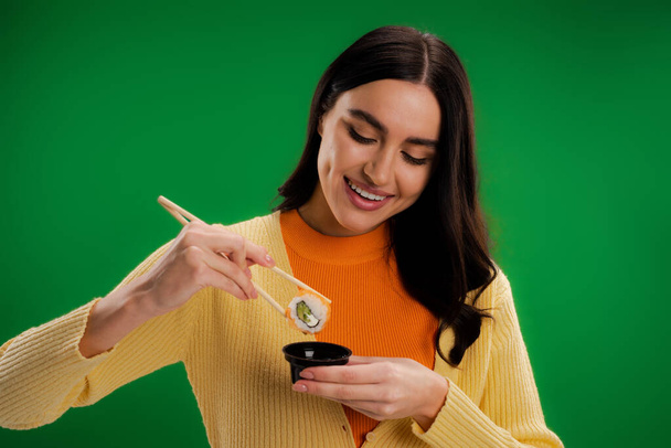 smiling brunette woman holding sushi roll near sauce bowl isolated on green - Fotografie, Obrázek