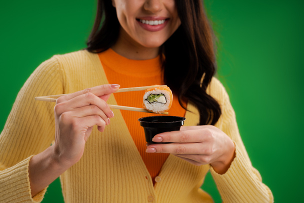 обрезанный вид улыбающейся женщины, держащей рулон суши рядом с соусом миска изолированы на зеленый - Фото, изображение