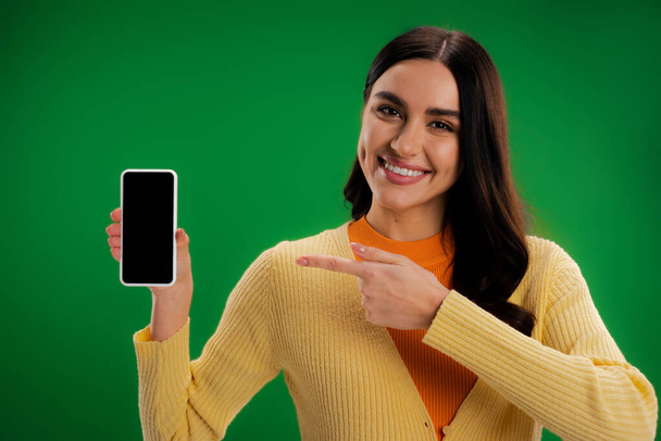 笑顔のブルネットの女性が緑色に隔離された空白の画面でスマートフォンを指して - 写真・画像