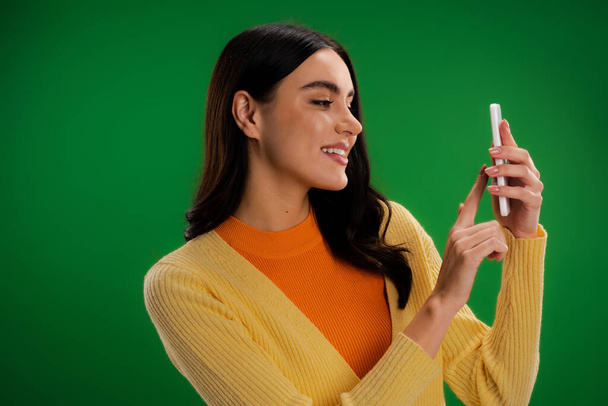 笑顔ブルネット女性で黄色ジャンパーとオレンジturtleeckで携帯電話を使用して隔離された緑 - 写真・画像