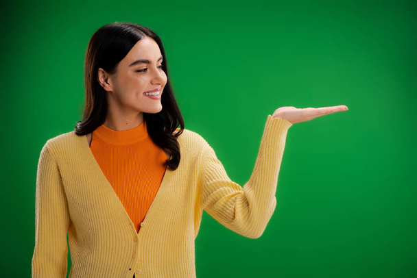 smiling woman in orange turtleneck and yellow jumper pointing with hand isolated on green - Φωτογραφία, εικόνα