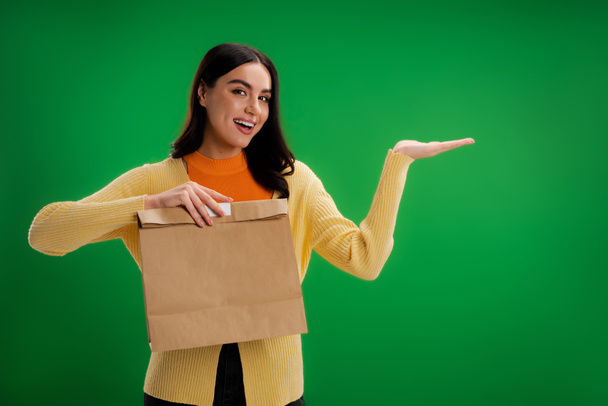 pleased woman holding food package and pointing with hand isolated on green - Foto, Imagen