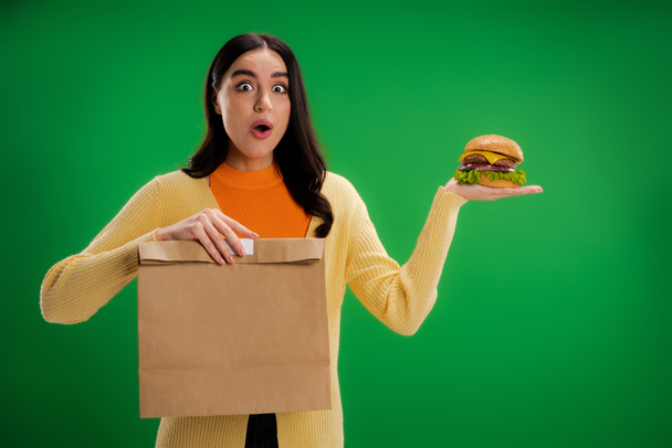 изумленная женщина с вкусным бургером и бумажным пакетом, глядя на камеру, изолированную на зеленый - Фото, изображение