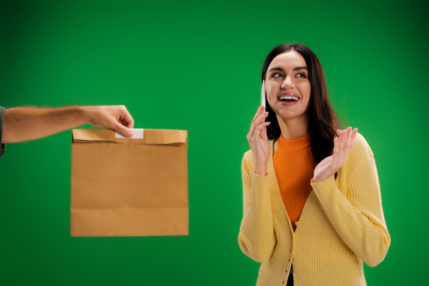 веселая молодая женщина звонит по мобильному телефону рядом с мужчиной с пакетом продуктов питания изолированы на зеленый - Фото, изображение