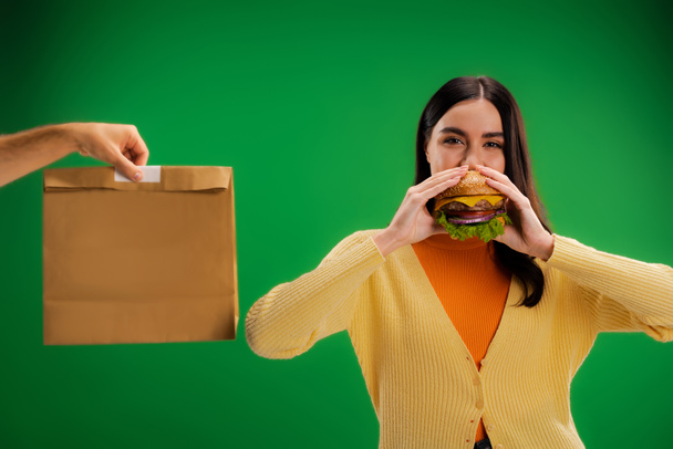 hungry woman eating delicious burger near man with food package isolated on green - Foto, immagini