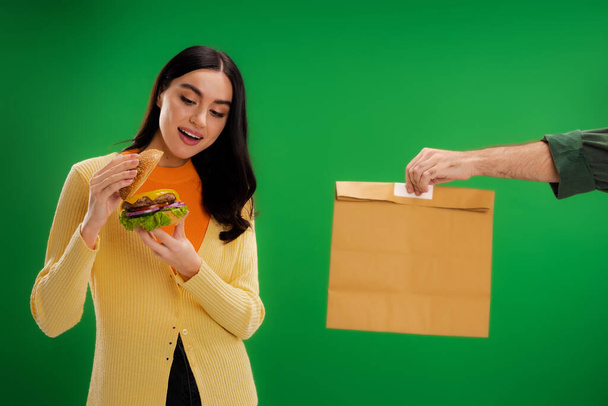 довольная женщина смотрит на вкусный бургер рядом с доставщиком мужчина с бумажным пакетом изолированы на зеленый - Фото, изображение