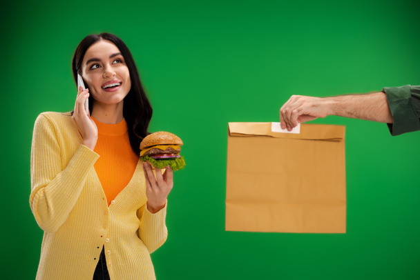 smiling woman holding burger and talking on smartphone near courier with paper bag isolated on green - Photo, Image