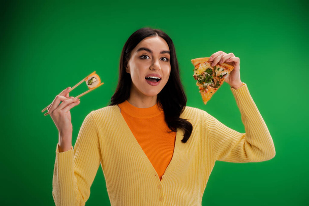 вражена жінка, дивлячись на камеру, тримає смачну піцу і свіжий суші-рол ізольовано на зеленому
 - Фото, зображення