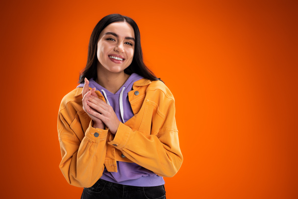 happy brunette woman in trendy jacket and purple hoodie looking at camera isolated on orange - Photo, image