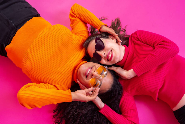 Two multi-ethnic women in sunglasses lying down laughing on a pink background, lifestyle - Foto, Imagen