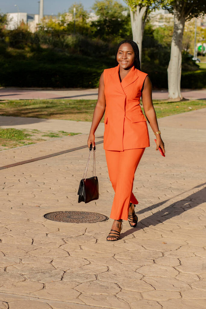 現代アフリカ系アメリカ人女性で鮮やかなオレンジ色のスーツチェック携帯電話ながら日の光の通りに立って - 写真・画像