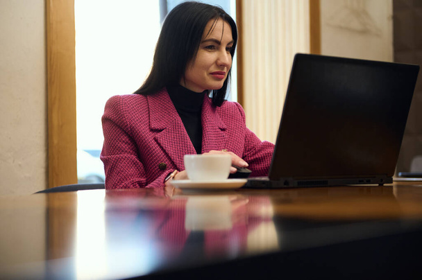 Successful Caucasian middle-aged woman entrepreneur, business person in casual wear, sitting at table with laptop and cup of coffee, while an online meeting with business partners, in office interior - Zdjęcie, obraz
