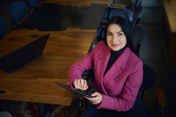 Attractive purposeful middle aged businesswoman, CEO, holding digital tablet, smiling cutely looking at camera, looking forward to meeting with business partners and investors at the negotiation table - Zdjęcie, obraz