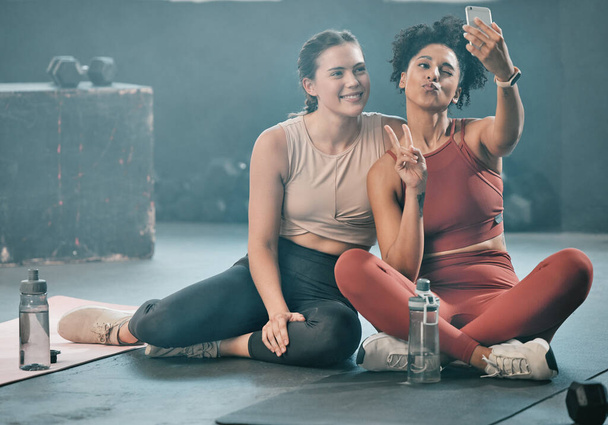 Woman, friends and fitness with phone for selfie, peace sign and pout after workout, exercise or training at gym. Sporty women with smile looking at smartphone for photo or healthy wellness together. - Photo, Image