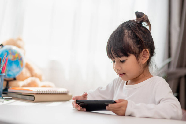 Asijské holčička pomocí telefonu zblízka, rozptýlení od studia, sedí u stolu s notebooky, hezké dítě baví s chytrým telefonem, sledování webinář, domácí výuka - Fotografie, Obrázek