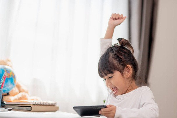 Azjatka korzystająca z telefonu z bliska, rozkojarzona z nauką, siedząca przy stole z notatnikami, śliczne dziecko bawiące się smartfonem, oglądające webinarium, uczące się w domu - Zdjęcie, obraz