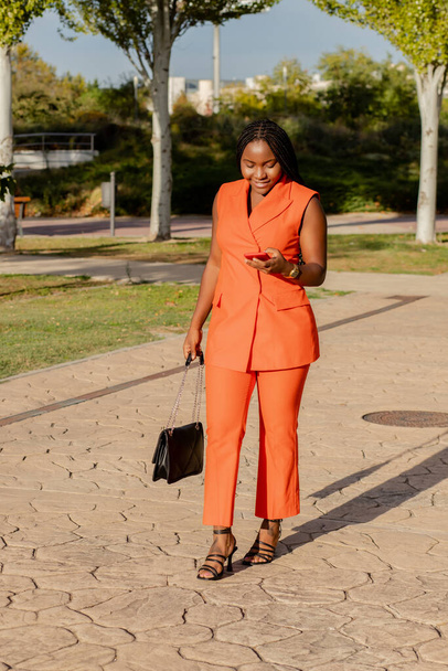Modern African American lady in vivid orange suit checking mobile phone while standing on sunlit street - Foto, imagen